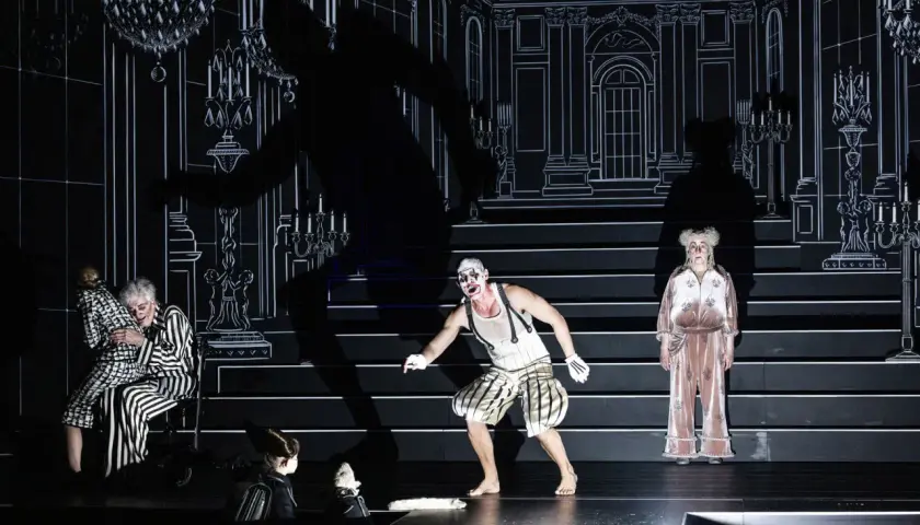 Scen från "Misantropen" på Dramaten. En clownliknande figur, en kvinna i randigt och en äldre kvinna i rosa pyjamas.