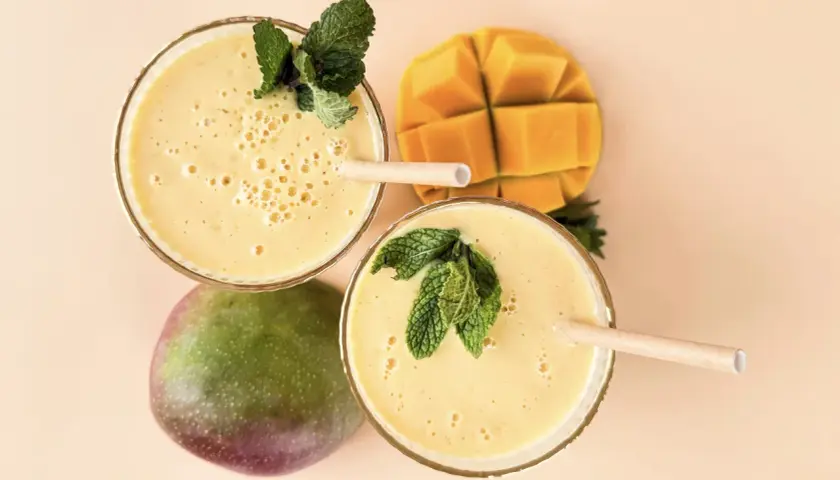 Zwei Gläser cremiger, veganer Mango Lassi, garniert mit Minze, neben einer ganzen und einer gewürfelten Mango.