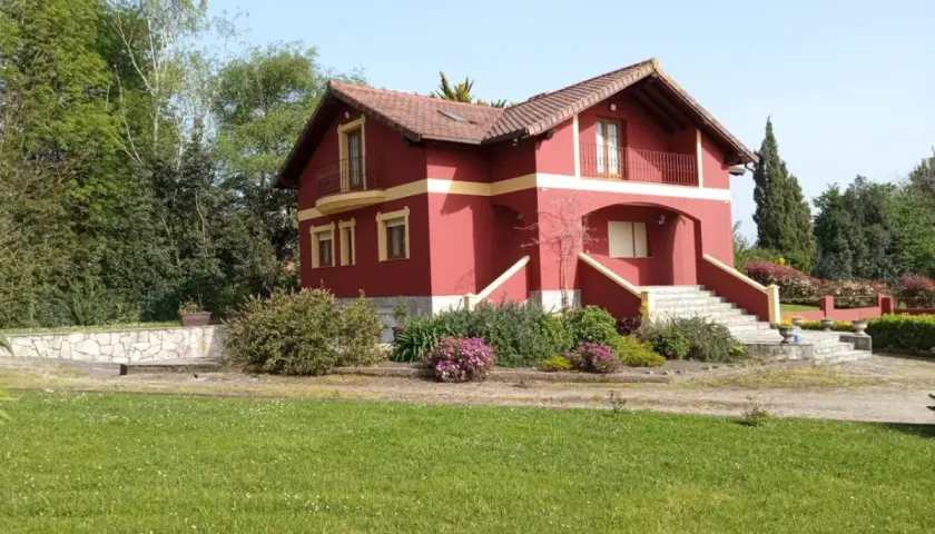 Una casa típica cántabra, de color rojo intenso con detalles amarillos, rodeada de vegetación y un césped verde.