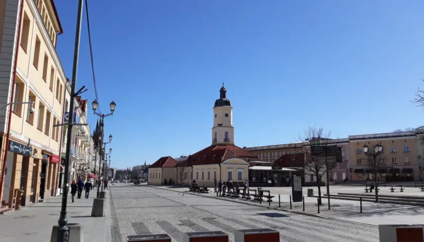 Słoneczny dzień na Rynku Kościuszki w Białymstoku. W oddali wieża ratusza, po bokach kamienice i brukowana nawierzchnia.