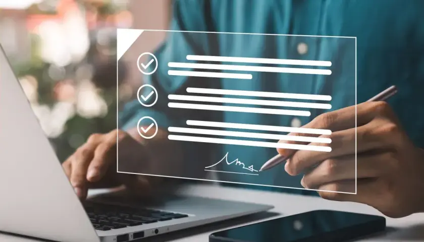 Dłoń podpisująca cyfrowy dokument z listą zadań na ekranie, obok laptop i smartfon.