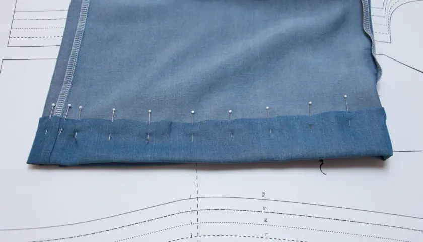 Pins secure the folded hem of blue knit pants, ready for sewing. This image shows a step in how to hem knit pants.