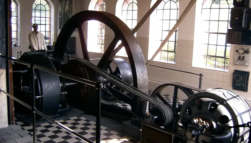 Eine historische Dampfmaschine, Symbol der Industrialisierung, mit großen Schwungrädern und Riemenantrieb. Ein Mann in Arbeitskleidung steht daneben.