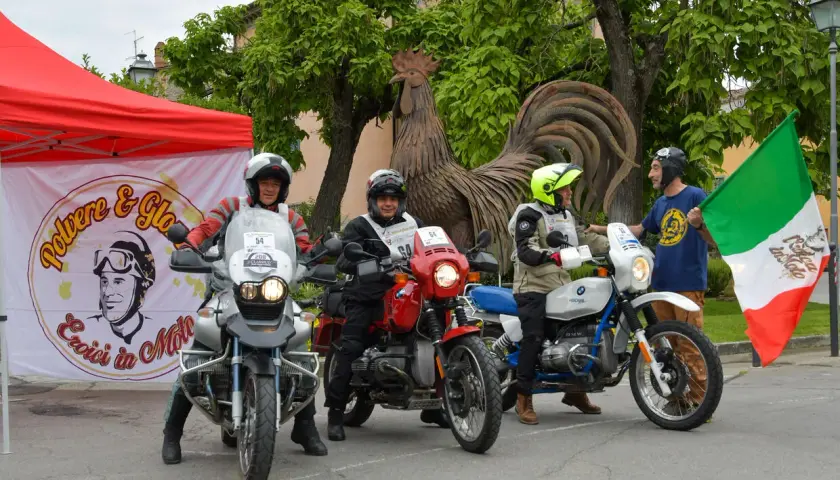 Tre motociclisti pronti per un percorso eroica in moto, con bandiera italiana e un grande gallo di metallo sullo sfondo.