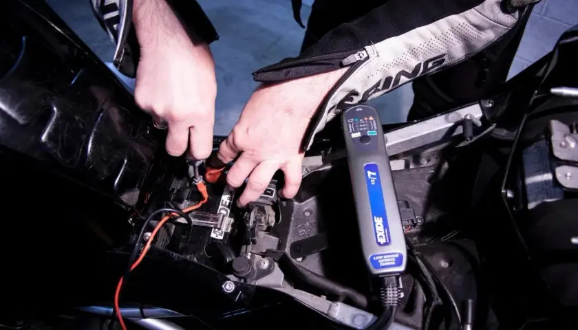 Mani che collegano un caricabatteria Exide alla batteria di una moto. Scopri come caricare batteria moto senza caricabatteria.