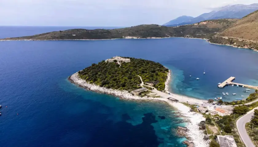 Isla con un castillo en Himare, Albania, rodeada de aguas azules y vegetación exuberante. Un muelle y barcos anclados completan la vista.