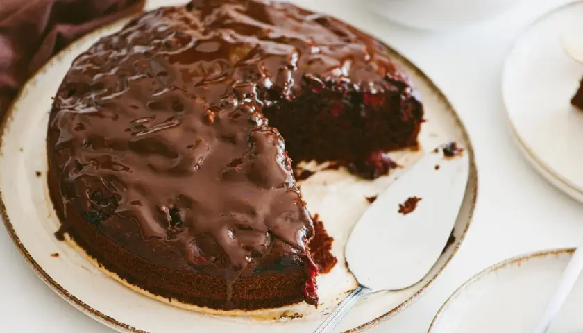 Pyszny brownie z malinami, polane błyszczącą czekoladą, czeka na talerzu. Obok stoi łopatka do ciasta.