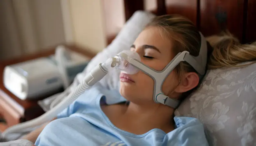Junge Frau mit Schlafmaske, die gegen Schlafapnoe-Symptome hilft. Ein CPAP-Gerät ist im Hintergrund zu sehen.