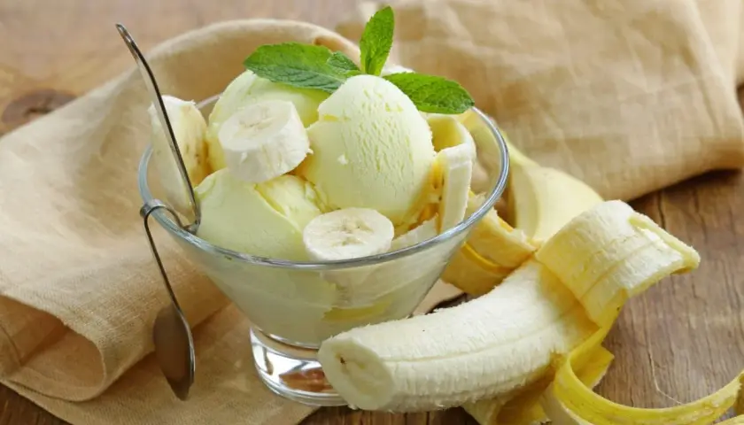 Pyszne lody bananowe w pucharku, ozdobione plasterkami banana i listkiem mięty. Obok leży obrany banan.