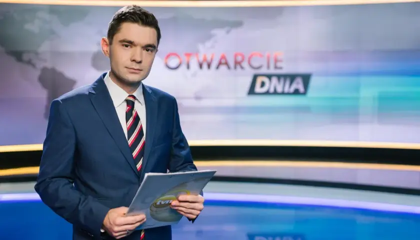 Jan Niedziałek, dziennikarz w garniturze, trzyma dokumenty w studiu telewizyjnym z napisem "OTWARCIE DNIA".