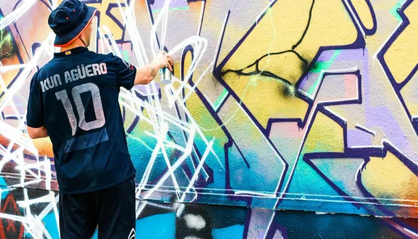 Mann mit Hut und Trikot sprüht mit Spraydose auf bunte Graffiti-Wand.