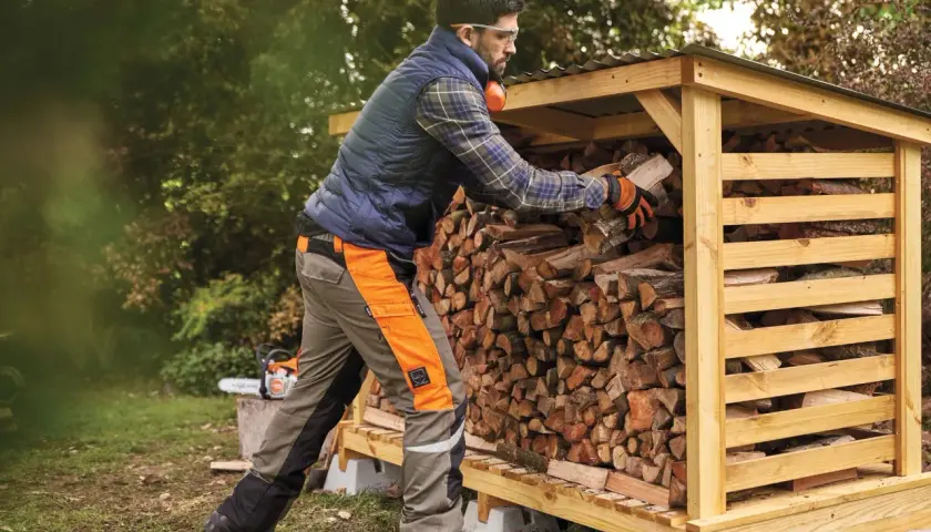 Mann stapelt Holz in einem Holzunterstand. Die Genehmigung für den Holzunterstand ist erteilt, und das Brennholz wird für den Winter vorbereitet.