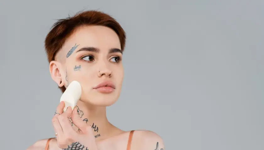 En kvinna med tatueringar applicerar foundation med en svamp för att jämna ut sin underton hud.