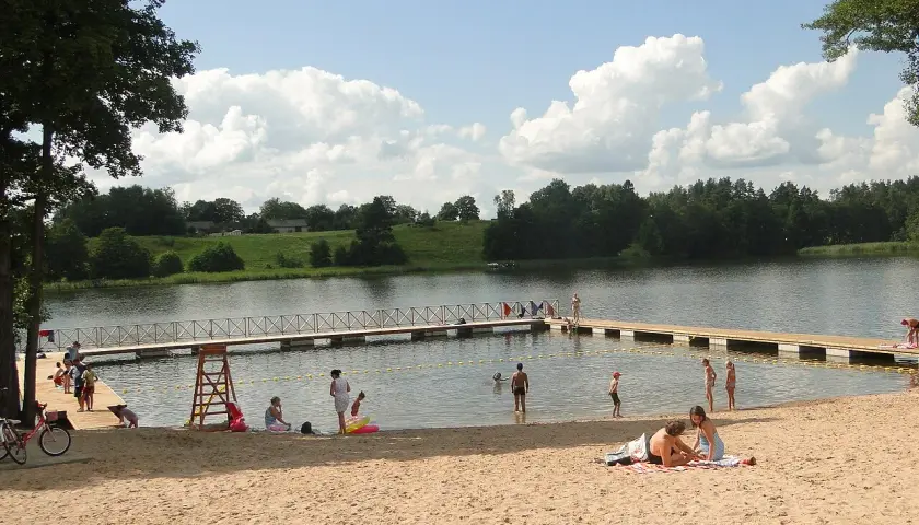 Słoneczne plaże na Mazurach tętnią życiem. Ludzie odpoczywają na piasku, kąpią się w jeziorze i spacerują po pomoście.