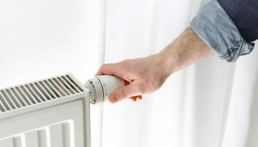 Dłoń reguluje termostat grzejnika, próbując obniżyć zbyt wysoką temperaturę w domu.
