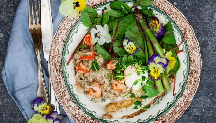 A plate of creamy risotto with shrimp, asparagus, greens, and edible flowers, served with a fork and knife.