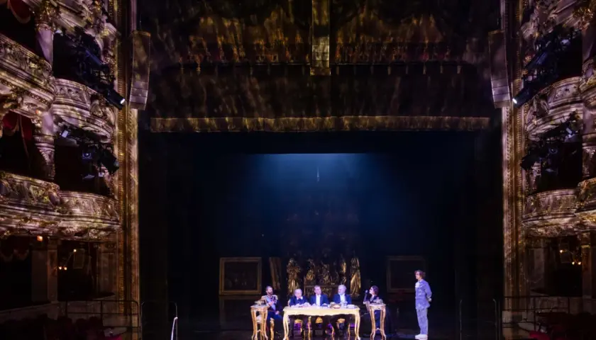 Na scenie teatru, przy złotym stole, siedzi pięć postaci. W tle widać bogato zdobione loże i kurtyny, tworząc atmosferę historycznego przedstawienia.