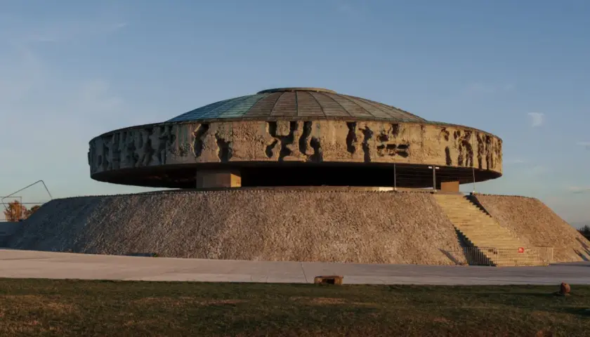 Monumentalne mauzoleum o okrągłej podstawie, z betonowym dyskiem ozdobionym płaskorzeźbami i szklanym dachem.