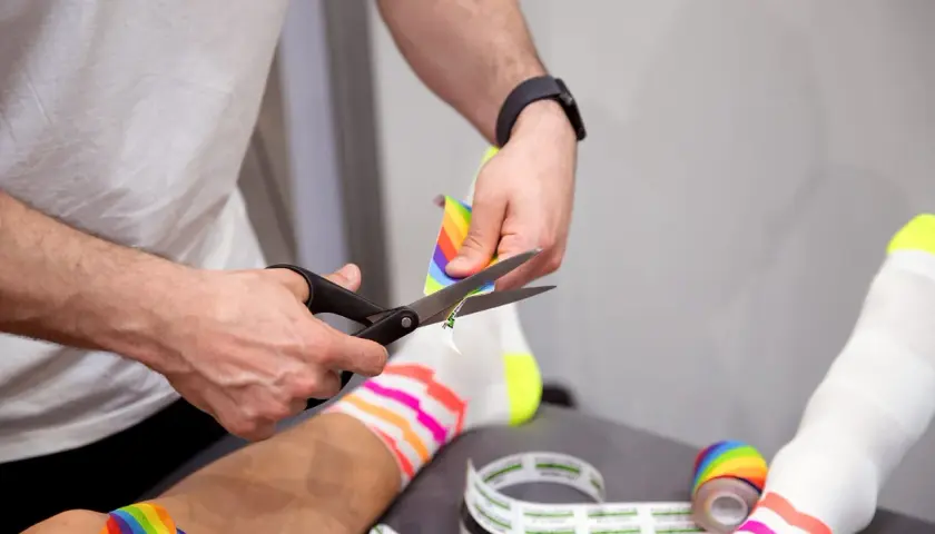 Hände schneiden buntes Kinesio-Tape zu. Die Frage "kinesio tape wann entfernen" wird hier visuell beantwortet: wenn es zugeschnitten wird, ist es bereit zur Anwendung.