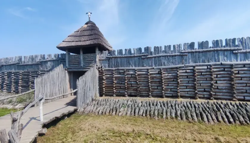 Drewniany gród obronny z wieżą i strzechą, pod niebieskim niebem.
