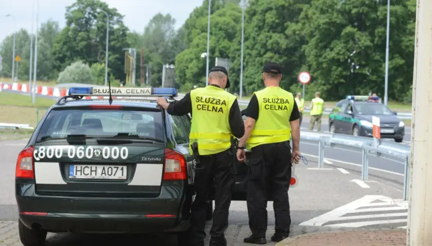 Służba celno skarbowa kontroluje ruch na drodze. Dwóch funkcjonariuszy w kamizelkach odblaskowych stoi przy oznakowanym radiowozie.