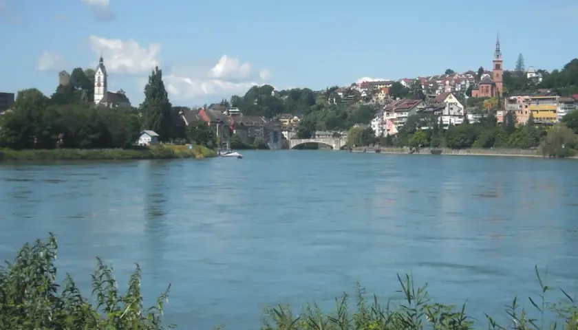 Un río azul fluye hacia una ciudad con iglesias y casas en la ladera, parte de la ruta EuroVelo 15.