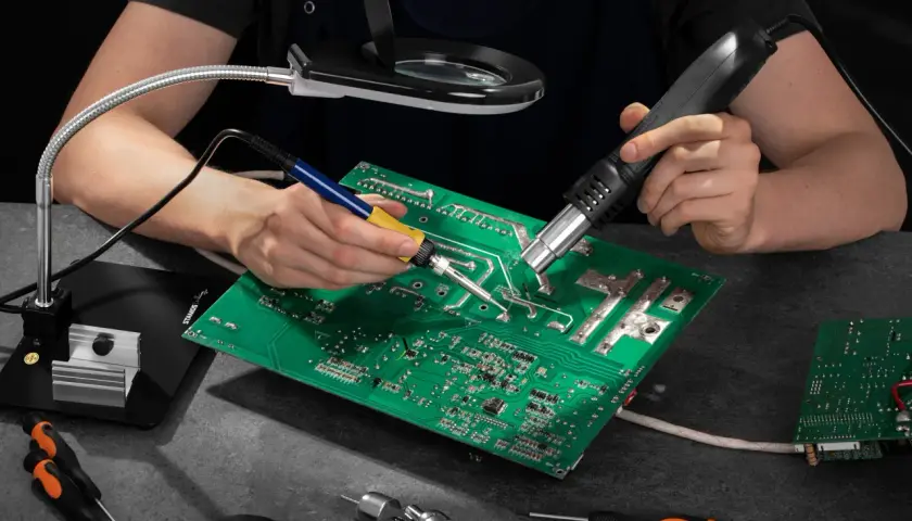 Ręce lutują płytę elektroniczną, używając lutownicy i gorącego powietrza. Widać też lupę, śrubokręty i taśmę izolacyjną – wszystko, co można lutować.