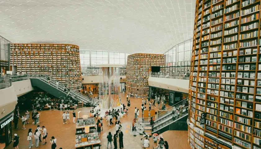 Wielopoziomowa biblioteka z półkami sięgającymi sufitu, gdzie każdy **bibliotekarz** marzyłby o pracy. Ludzie spacerują, czytają i odpoczywają wśród książek.