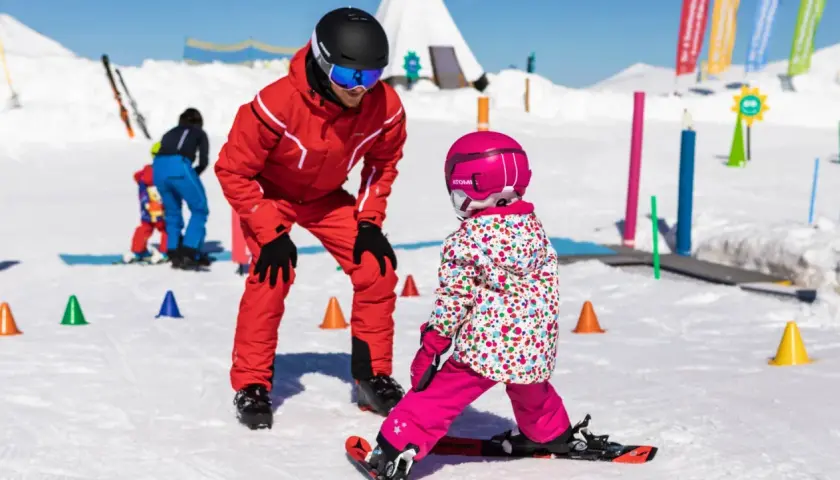 Maestro di sci insegna a bambini a sciare su pista innevata, con coni colorati e bandierine sullo sfondo.