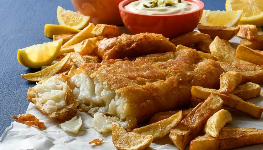 Un delicioso plato de fish and chips, con pescado rebozado crujiente, patatas fritas doradas, guisantes y salsa tártara.