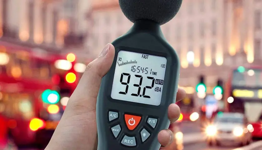 Medidor de sonido en mano, mostrando 93.2 dBA en una calle ruidosa, midiendo la potencia acústica del tráfico urbano.