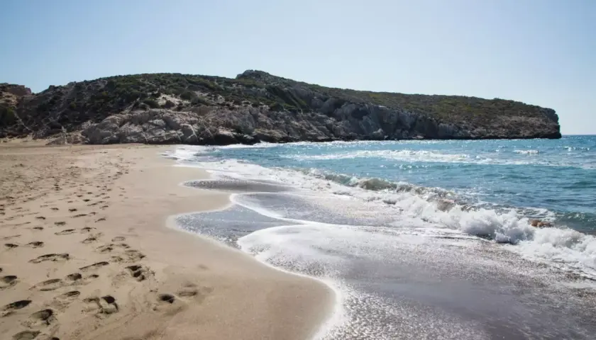 Piaszczysta plaża z falami i skalistym wzgórzem w tle.