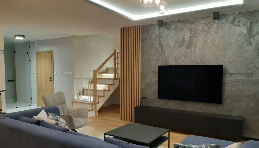 Nowoczesny salon z szarą ścianą i telewizorem. Drewniane lamele idealnie komponują się ze schodami i podłogą, tworząc ciepłą atmosferę.