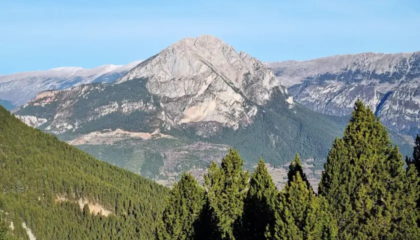 Majestuosa Serra d'Ensija bajo un cielo azul. Bosques de pinos cubren las laderas, con picos rocosos y nevados al fondo.