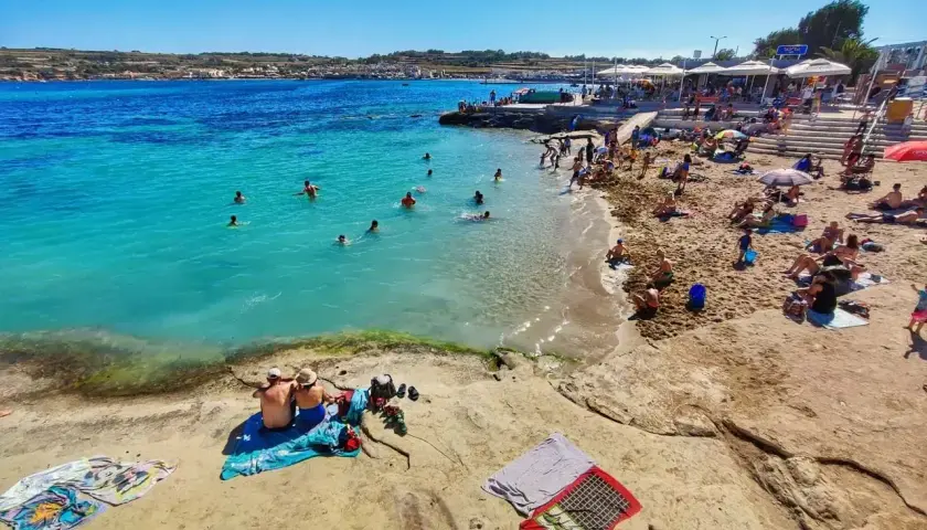 Plaża na Malcie z turystami kąpiącymi się w turkusowym morzu i odpoczywającymi na piasku.