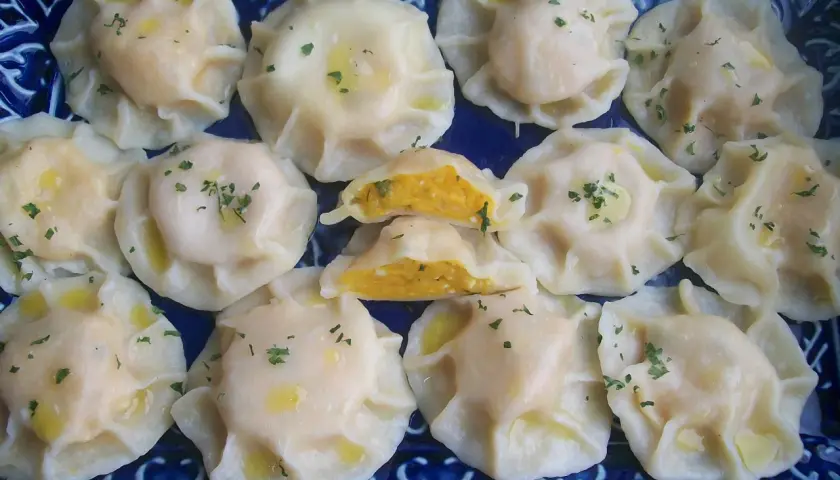 Pyszne pierogi z dynią, polane masłem i posypane natką pietruszki, ułożone na niebieskiej tacy.