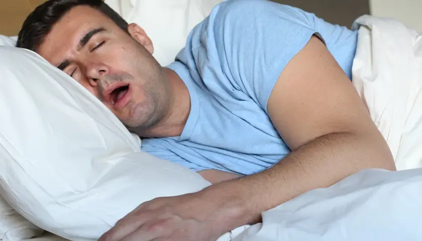 Uomo dorme a bocca aperta, spiegando perchè si sbava quando si dorme. La sua testa è appoggiata su un cuscino bianco.