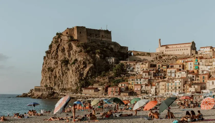 A sunny beach with people relaxing under umbrellas, overlooking a cliffside town with a castle. This view is a perfect destination for those wondering how to get to Calabria, Italy.