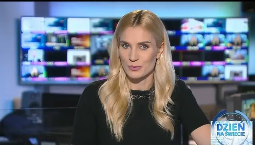 Blond dziennikarka TVN w czarnej bluzce z naszyjnikiem, na tle monitorów.
