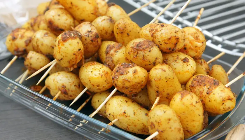 Des brochettes de pommes de terre assaisonnées, prêtes pour le barbecue, dans un plat en verre.