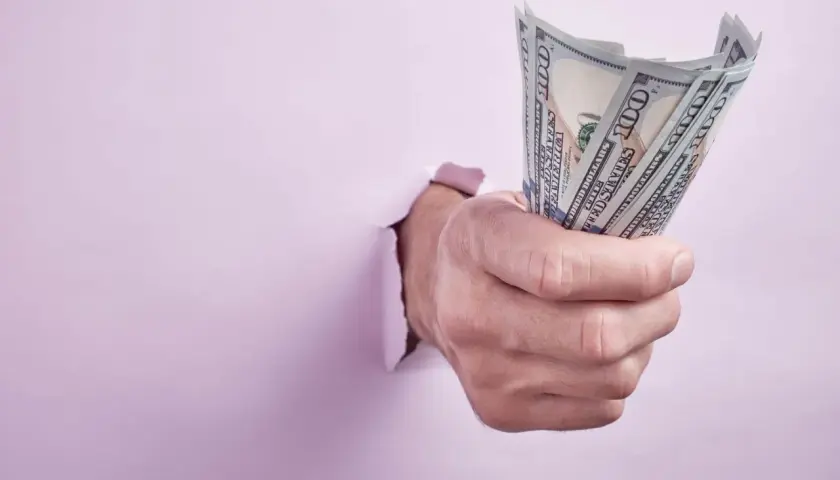 Dłoń przebija się przez różową ścianę, trzymając plik banknotów 100 dolarowych. Może to symbolizować zwrot kaucji od byłego lokatora.
