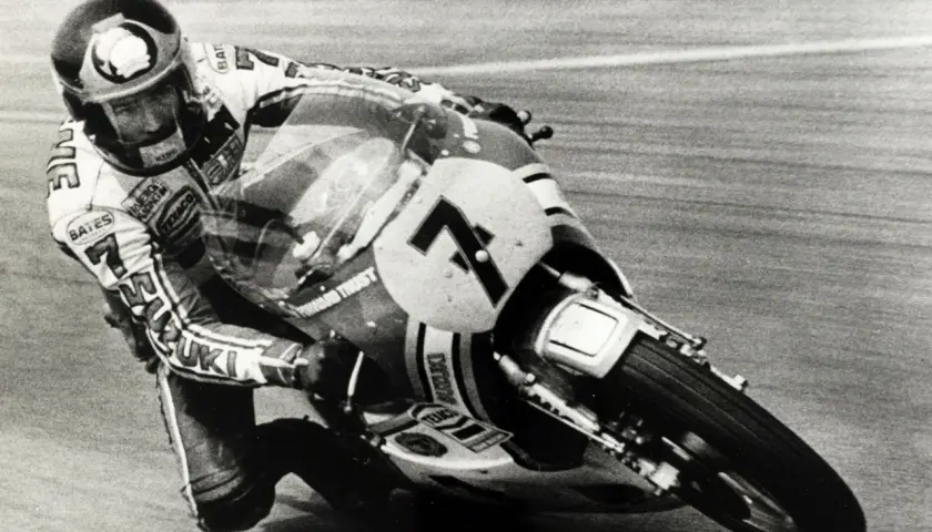 Wyścigowy motocykl Suzuki z numerem 7 pochylony w zakręcie, z zawodnikiem w kasku. To fragment historii motocykla.