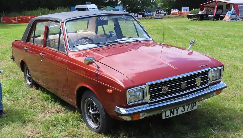Röd Hillman bil med svart vinyltak, parkerad på en gräsmatta vid ett evenemang.