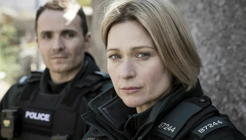 Två poliser i uniform, en kvinna i förgrunden med intensiv blick, bakgrunden svagt belyst av blåa ljus.