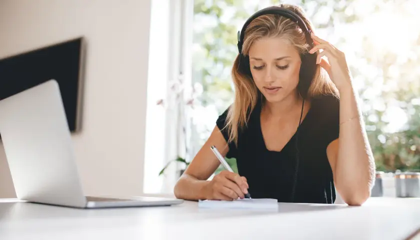Mujer con auriculares escribe en un cuaderno, con luz natural entrando por la ventana. La luz suave es ideal, preguntándose cuál es la mejor luz para estudiar.