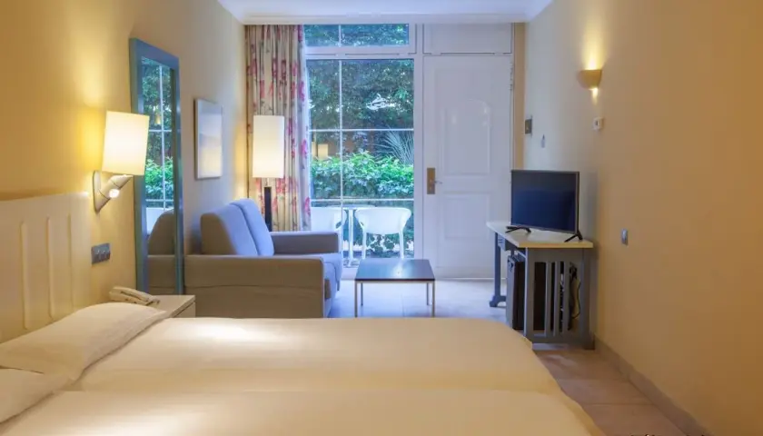 Pokój rodzinny w hotelu IFA Altamarena: łóżko, sofa, TV i wyjście na taras z widokiem na zieleń.