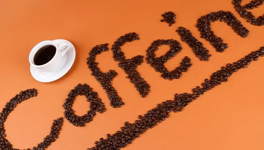 Ziarna kawy ułożone w napis "caffeine" obok filiżanki kawy. Zastanawiasz się, ile kofeiny ma łyżeczka kawy mielonej?