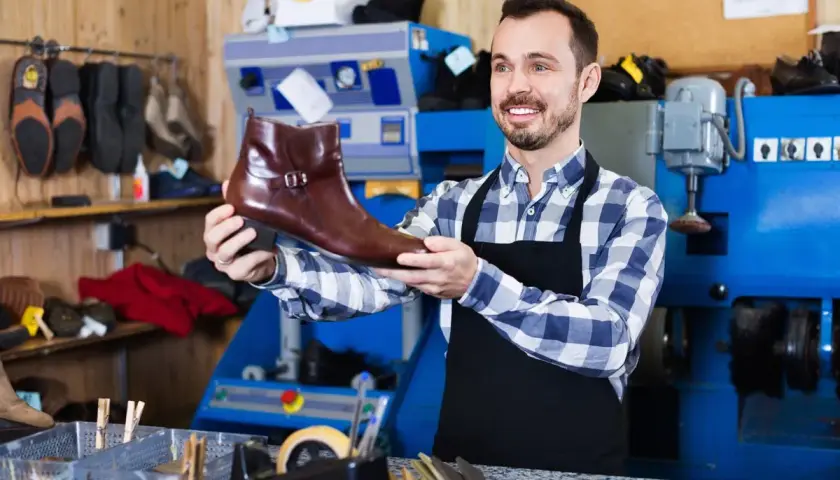 Artisan souriant tenant une botte en cuir marron, prêt pour la réparation de chaussure. L'atelier est bien équipé.