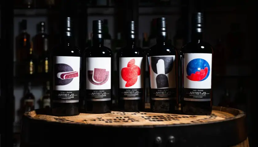 Cinq bouteilles de blend whisky Artist-13, ornées d'étiquettes artistiques colorées, reposent sur un fût de whisky.