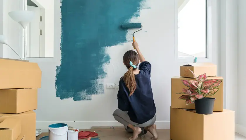 Mujer pintando una pared de azul, rodeada de cajas. Es la mejor época para pintar la casa y darle un nuevo aire.